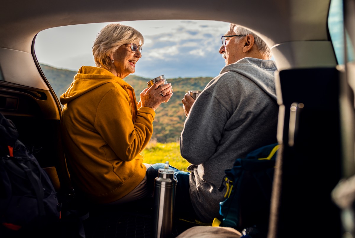 Senior couple sitting against the car, resting after hiking in c Senior couple sitting against the car, resting after hiking in countryside.